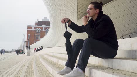 Side-view-of-a-young-man-looking-sad-and-frustrated-sitting-on-steps-wearing-a-black-jacket-and-glasses-with-an-umbrella