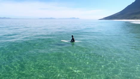 Vista-Aérea-De-Drones-De-Un-Tipo-Sentado-En-Su-Tabla-De-Surf-En-El-Agua-Esperando-Olas