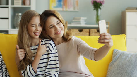 Portrait-shot-of-the-pretty-Caucasian-mother-and-her-teen-daughter-taking-selfies-photos-on-the-white-smartphone-in-the-modern-living-room.-Indoor.