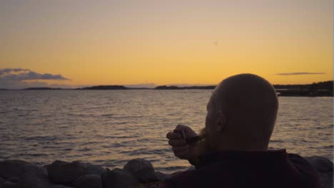 caucasian male with short hair and ginger beard is looking towards the better days sitting by the sea ocean on stony shore smoking his antique pipe releasing clouds of smoke while the sun is setting