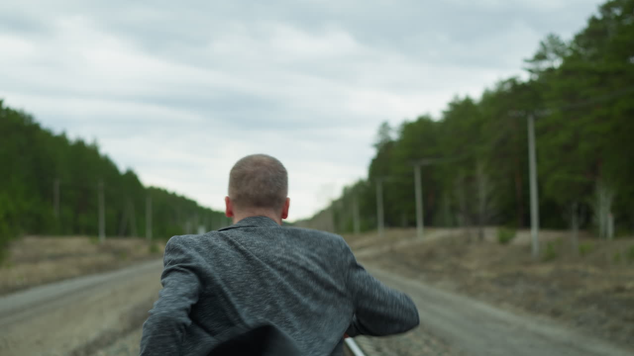 una vista cercana de un hombre anciano corriendo por una vía ferroviaria, vestido con un traje gris y vaqueros, la vía ferroviaria está cubierta de piedras, y con una vista borrosa de árboles y postes eléctricos