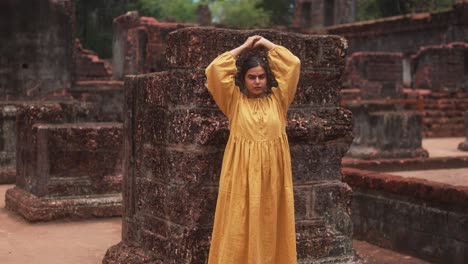 an indian woman in a yellow dress leans against a weathered stone pillar at an ancient historical site, india