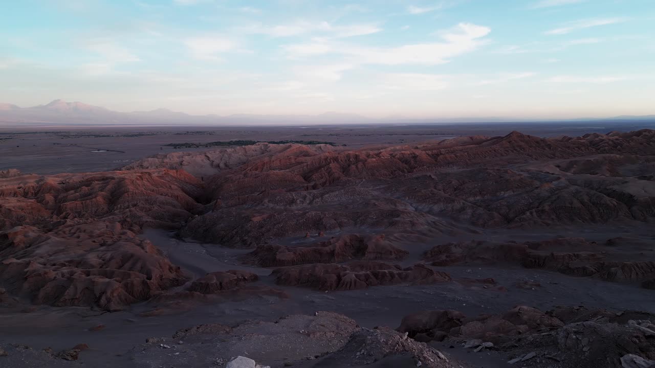 Vista panorâmica sobre dramáticas formações rochosas vermelhas erodidas do Vale da Lua ao anoitecer com a vasta planície do deserto do Atacama e vulcão distante no horizonte. San Pedro de Atacama, Chile