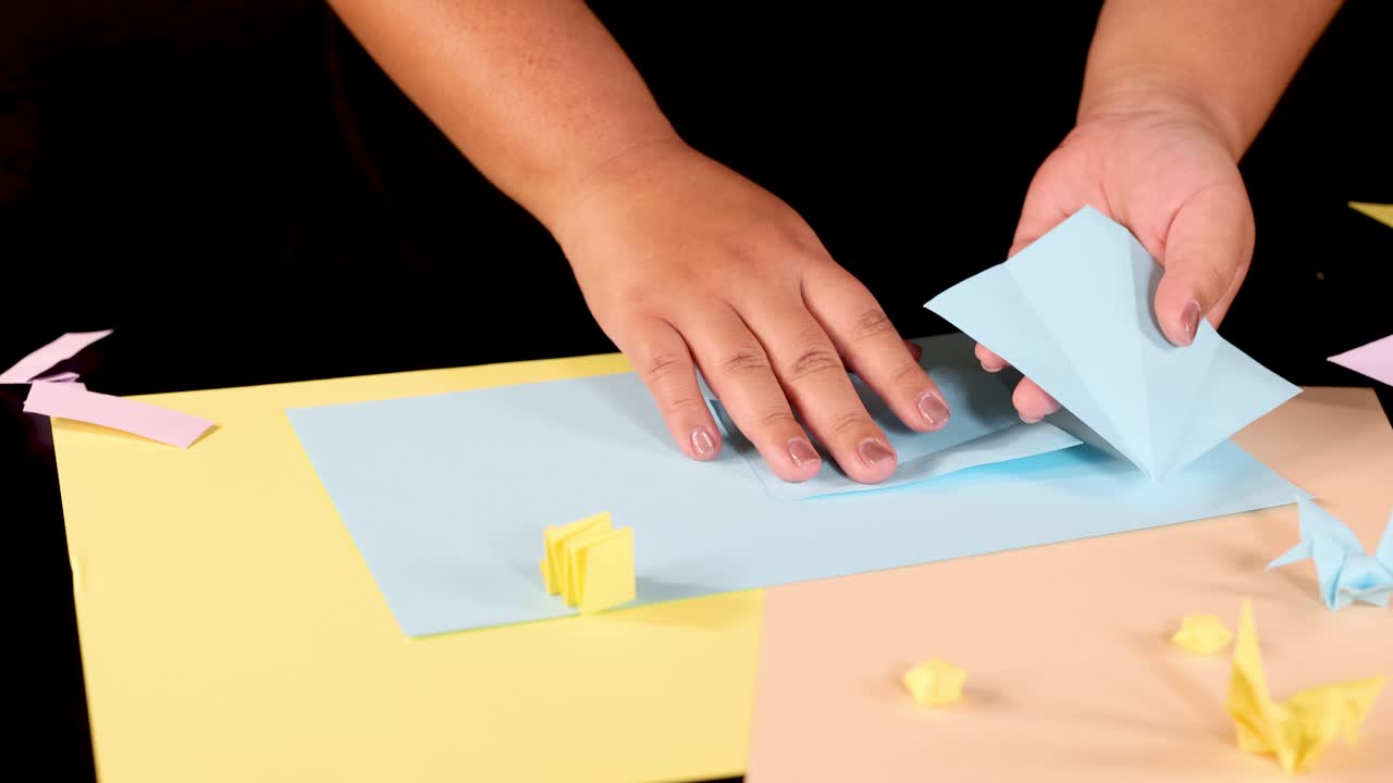 Person folds blue paper into origami shape on pastel sheets under bright, even lighting