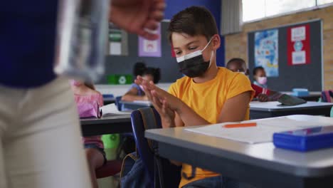 Mixed-race-schoolboy-wearing-face-mask-disinfecting-hands-in-classroom,-colleagues-in-background