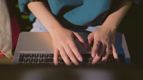 close-up of woman's hands typing on laptop outdoors at night, screen reflection illuminates her fingers while she works on bench with red bag nearby