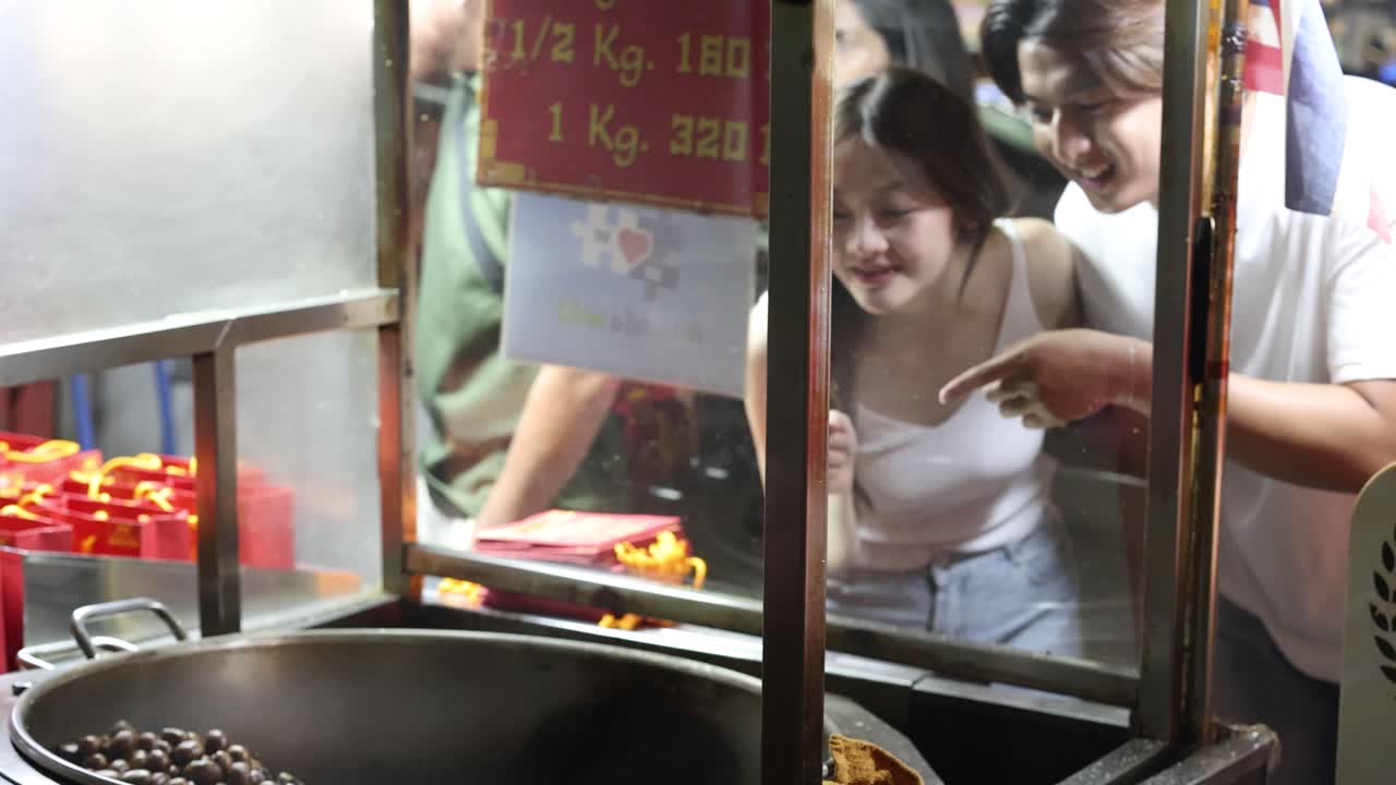 Smiling couple selects roasted chestnuts at brightly lit night market vendor stall, lively urban atmosphere