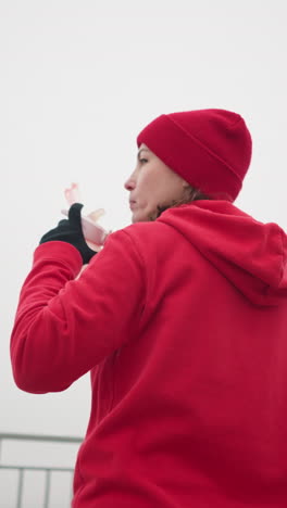 back view of woman in red hoodie and beanie sipping water outdoors with pink bottle, drops it down, foggy atmosphere, blurred background of iron railing, misty scenery, serene winter outdoor moment