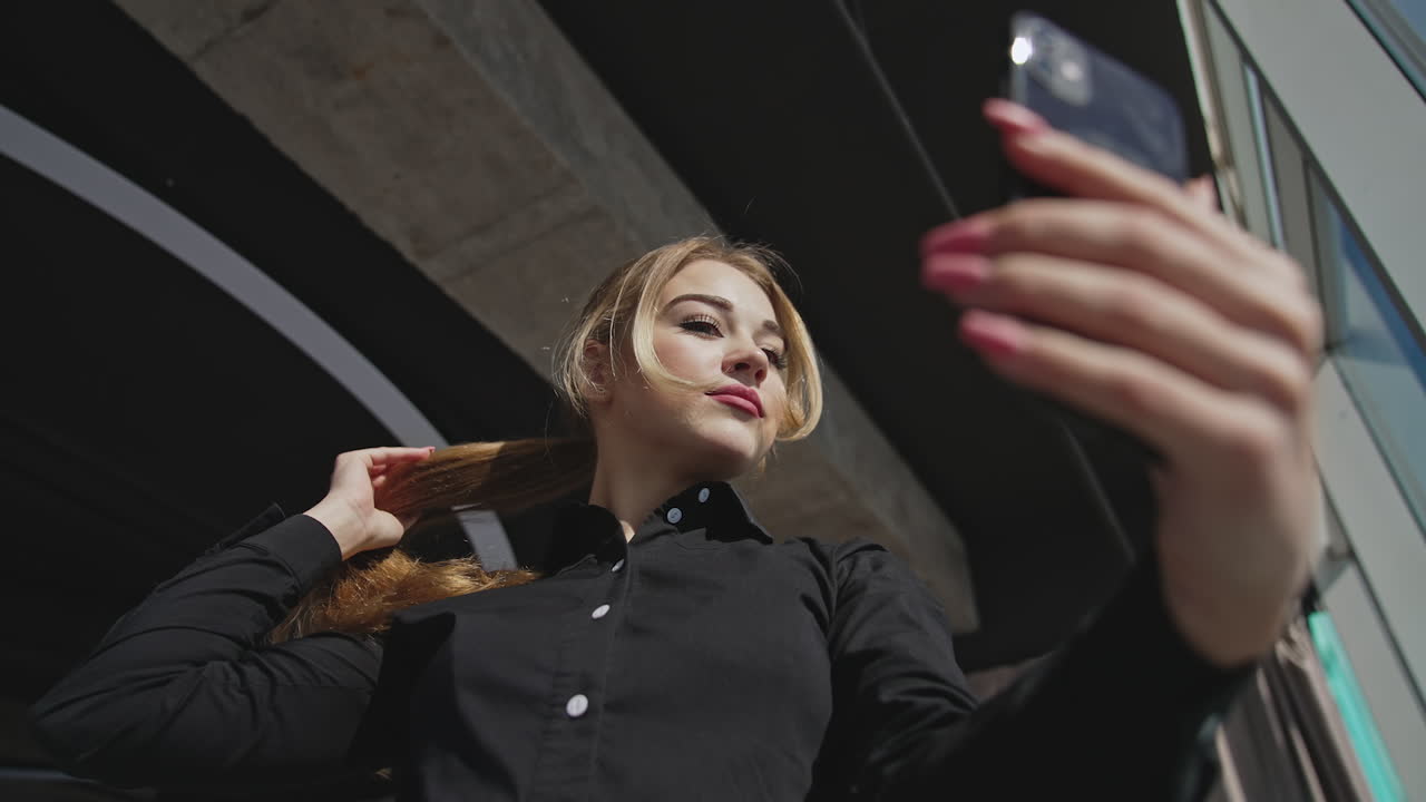 Attractive lady trying to find the best pose for taking selfie. Young woman changing postures standing at the big panoramic window. Low angle view.