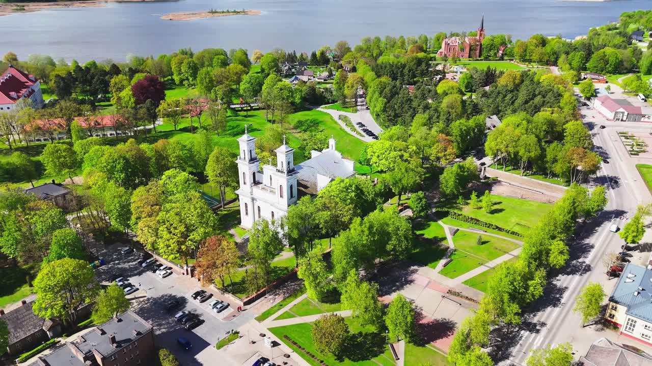 Twin white church towers stand in a vibrant green park near the river, facing a red brick cathedral across a historic European town under a bright spring sky.