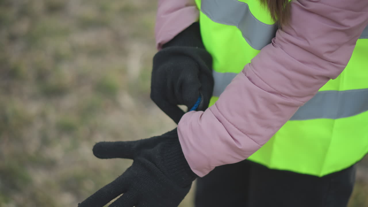 Close-up of person in high-visibility neon yellow safety vest with reflective stripes and pink padded jacket putting on black knitted gloves outdoors during cold season, preparing for safety work