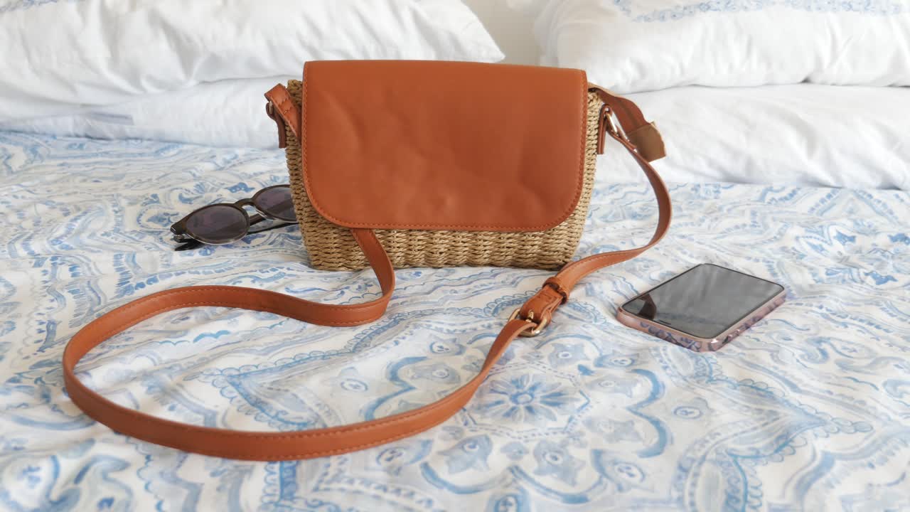 Borsa a tracolla in paglia marrone sul letto con telefono e occhiali da sole