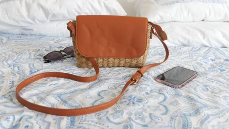 brown straw crossbody bag on bed with phone and sunglasses