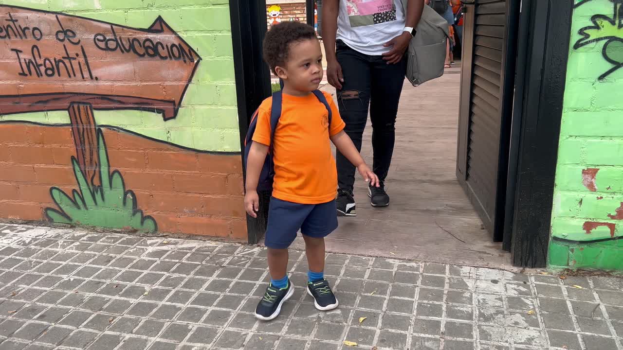adorable niño europeo africano de dos años que vive la enfermería después de su primer día después de las vacaciones de verano, vestido con una camisa naranja, pantalones cortos azules y una mochila, seguido de mamá