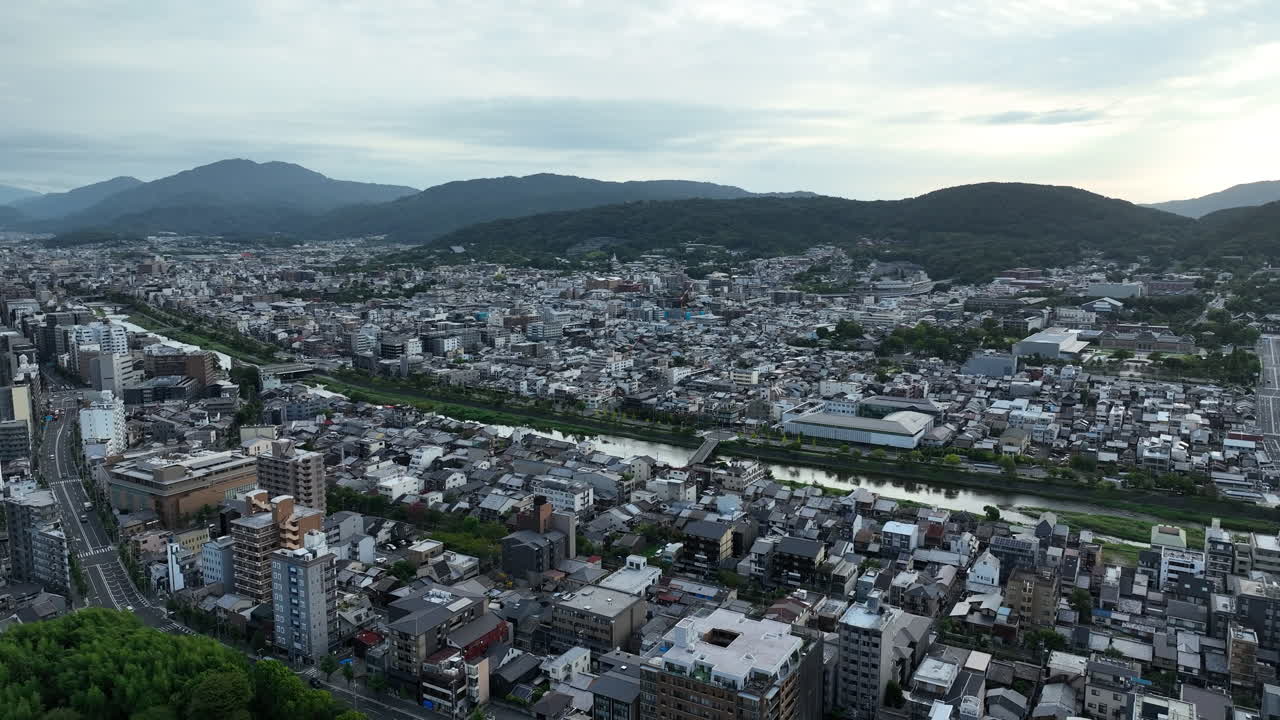 River Through Dense Buildings Backdropped By Scenic Hills In Kyoto, Japan. wide aerial shot