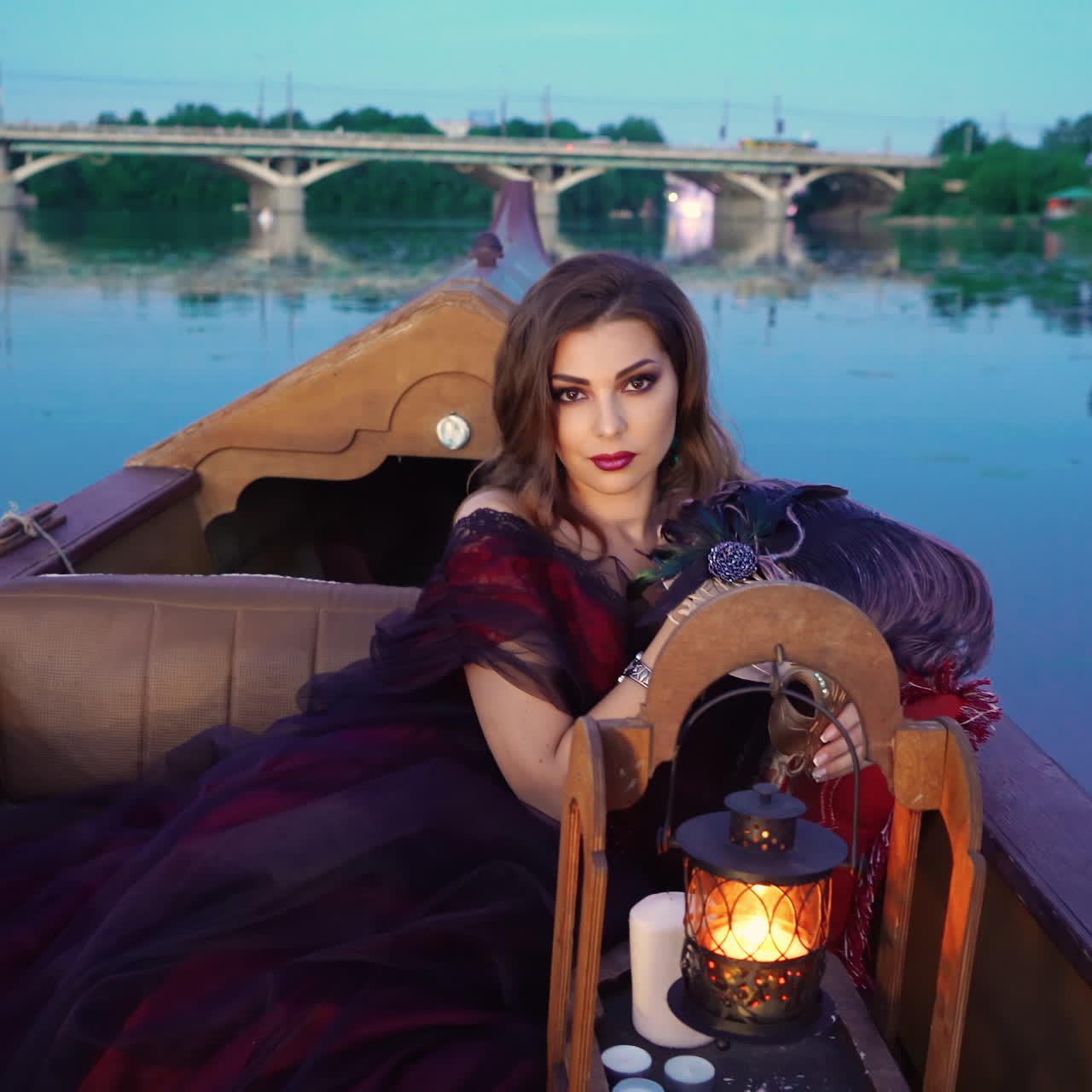 Lovely sexy woman in evening dress sitting in a boat and looking at camera with passion on the river with a big bridge. Pretty female floating in the evening