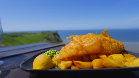 traditional british fish and chips overlooking coastal sea beach landscape in background on summer day in wales. illustration of fresh seafood from boat to plate. served with chips, peas, lemon.
