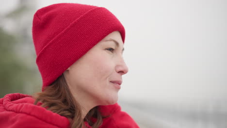 close-up of woman in red hoodie and beanie gazing thoughtfully into distance with soft smile, set in a misty outdoor atmosphere creating a serene and introspective winter mood