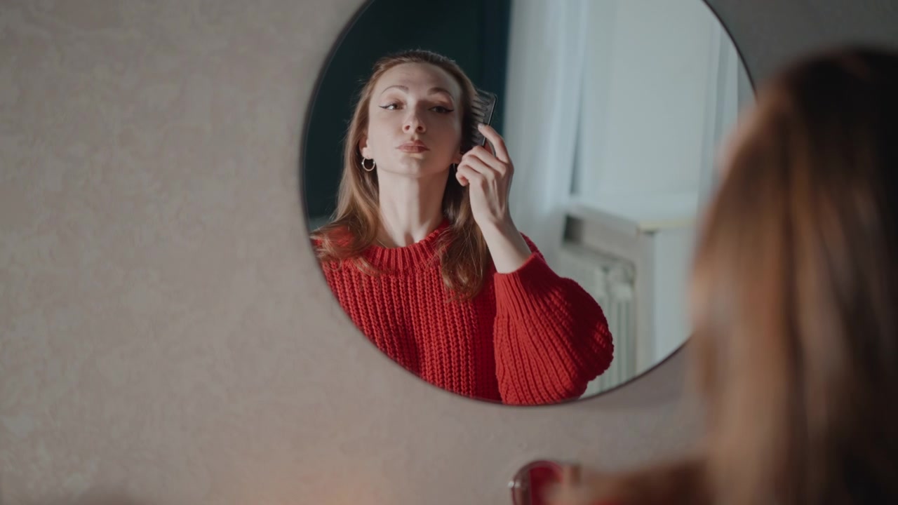 Donna si sistema e liscia i capelli mentre osserva il suo riflesso in uno specchio rotondo a parete all'interno di un'accogliente camera da letto, con un delicato movimento della mano, una leggera inclinazione della testa e una soffusa luce naturale.