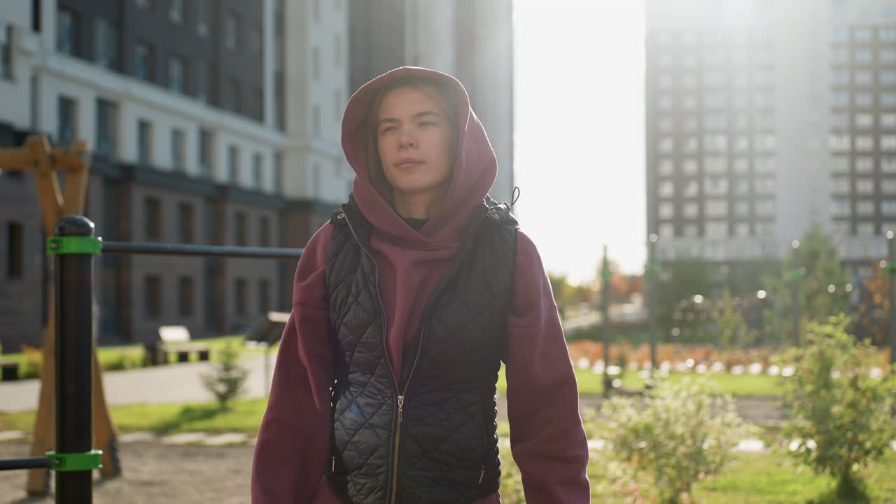 Gym goer in hoodie and vest stretches arms and legs outdoors in urban park with pedestrians passing by playground bars under morning sunlight preparing workout routine with focus and energy