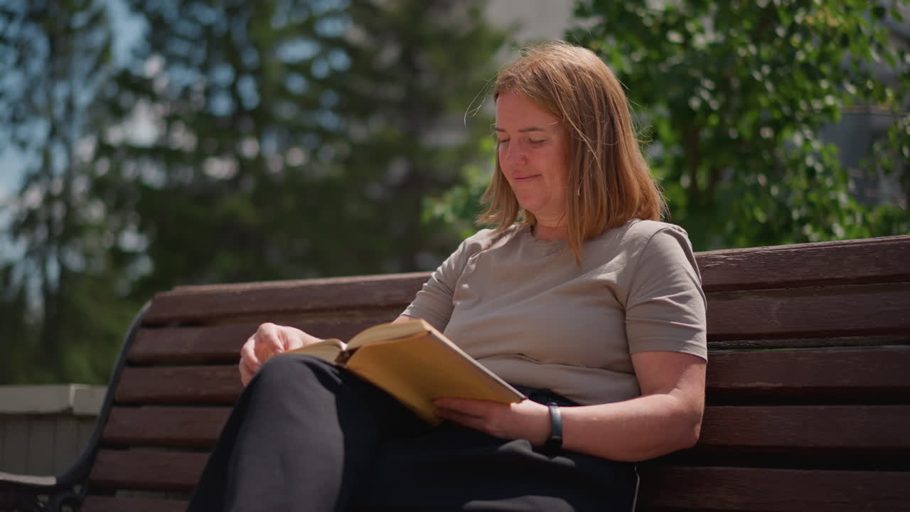 Une femme assise sur un banc en bois, lisant un livre à l’extérieur sous une lumière vive, expression détendue et posture calme, voitures floues passant à l’arrière-plan, mélangeant étude paisible.