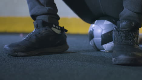 close-up of black athletic shoes on turf next to a soccer ball under soft indoor lighting, detail of footwear and sports equipment, with yellow panel in the background