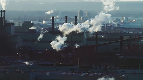 industrial complex with smokestacks emitting steam and smoke, city backdrop, early morning light