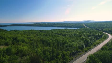 smooth drone shot over the trees and hydro lines revealing a highway next to the shimmering blue water of eastern canada