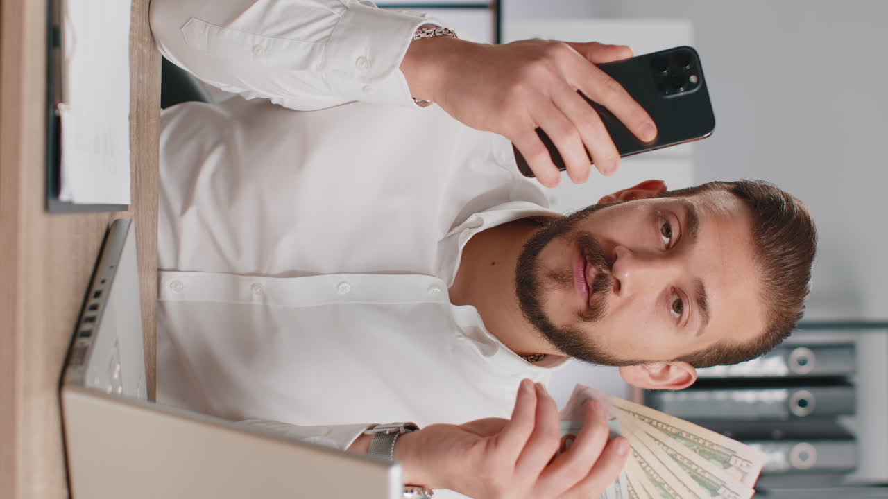 Uomo d'affari che mostra lo smartphone, si rallegra sinceramente per una vittoria, riceve denaro, dollari in contanti, banconote.