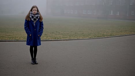 young student standing in a foggy schoolyard during a cold morning, wearing a blue coat and a scarf, with a school building in the background
