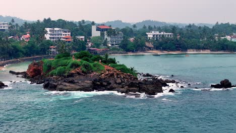 sri lanka lush rocky islet in turquoise bay near palm-fringed coast town. waves break on rocks under hazy sky. elevated viewpoint captures tropical scene
