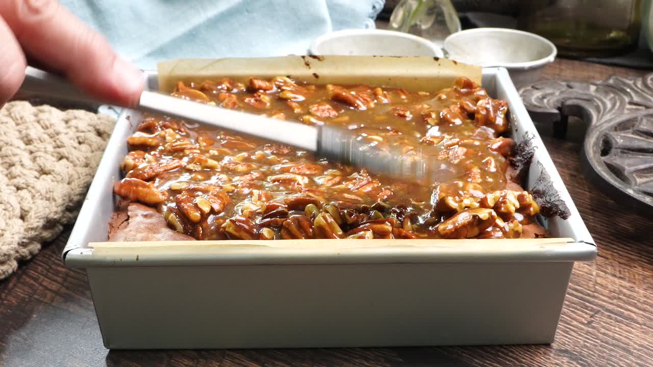 Baking delicious caramel pecan brownie. Using a spatula to smooth out the layer of caramel and pecan on top of the brownie. Freshly baked gooey chocolate brownies with sweet caramel and nutty pecans.