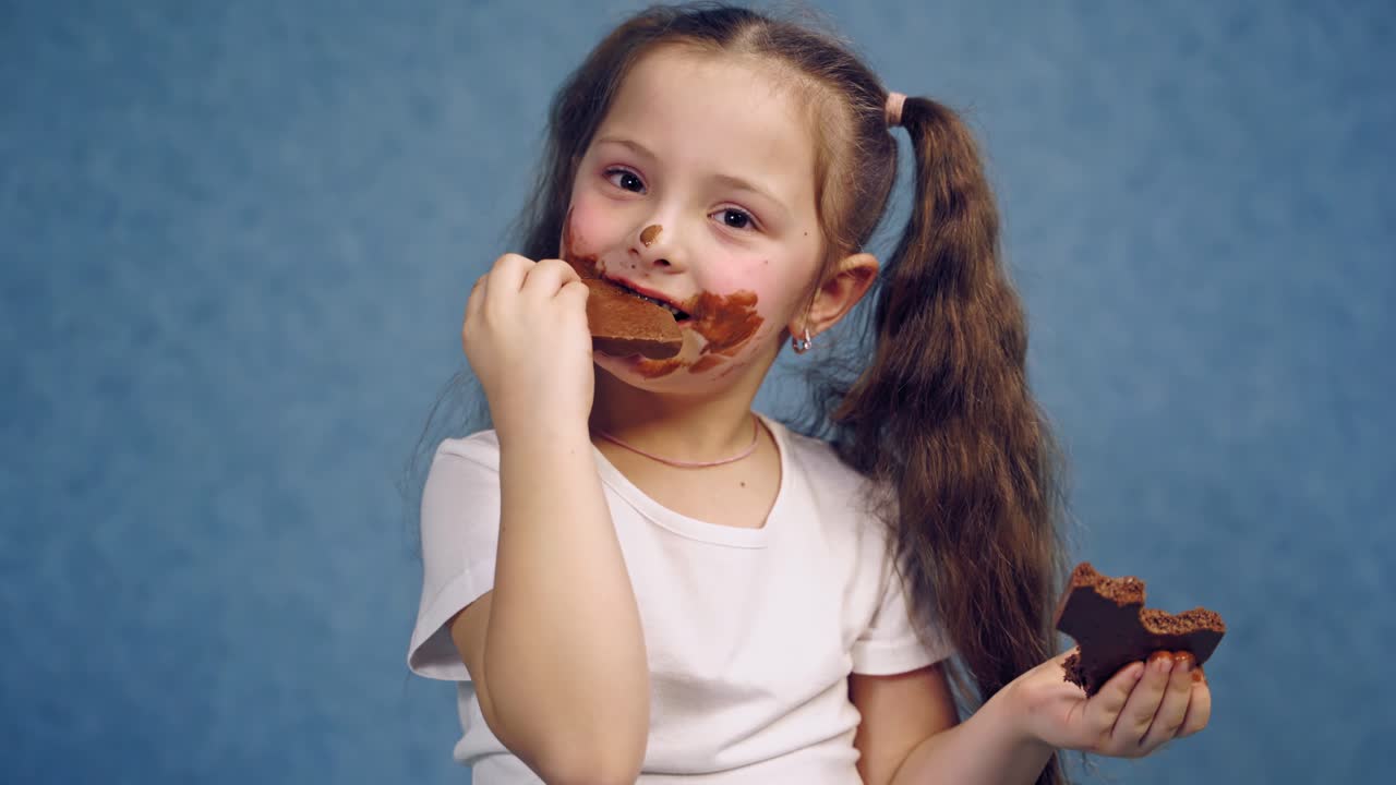Little girl with dirty face eats bar of chocolate. Funny child with two ponytails is biting tasty black chocolate on blue studio background.