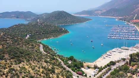 Drone-Aéreo-De-Una-Península-Llena-De-Colinas-Verdes-Y-Agua-Azul-Turquesa-Tropical-Con-Vistas-Al-Mar-Mediterráneo-En-Kas-Turquía-En-Un-Día-Soleado-De-Verano-Con-Un-Puerto-Deportivo-Y-Barcos-Atracados