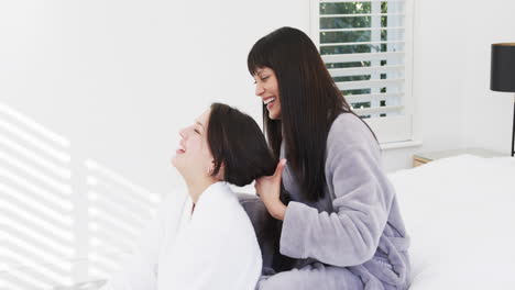 Happy-biracial-mother-in-bathrobe-brushing-hair-of-smiling-adult-daughter-in-sunny-room,-slow-motion