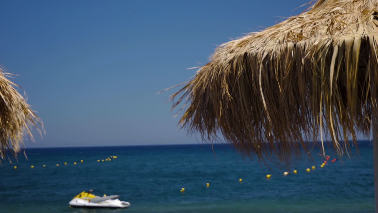 Handheld Medium Shot of a Tiki Hut Umbrella With Dried Palm Leaves on a Sunny Day in Perissa Santorini in Greece Europe With a Jetski and a Woman in Red in The Background