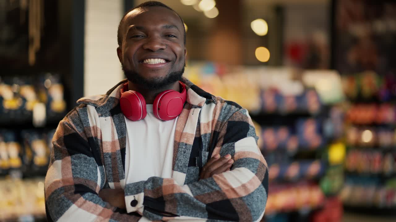 Portrait of a happy and confident man with Black skin color with a beard in a checkered shirt and red wireless headphones who crosses his arms on his chest miles and poses among the counters in a modern grocery store