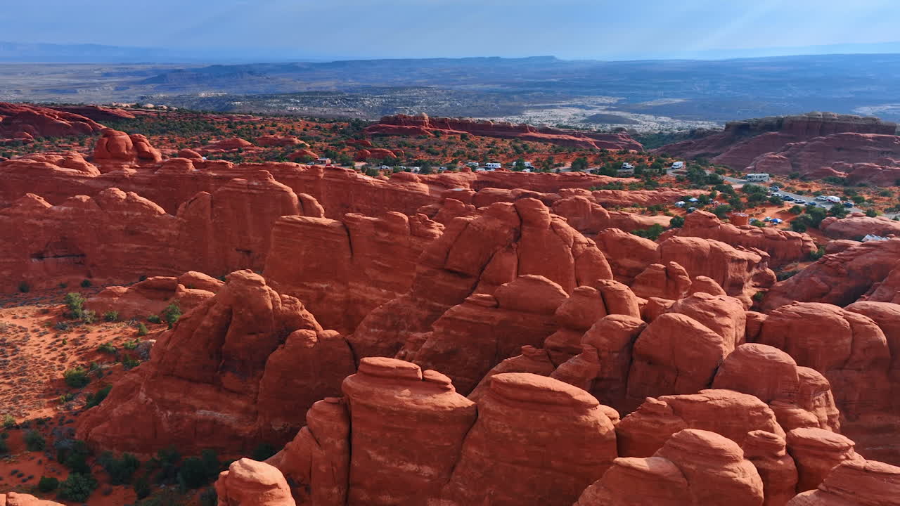 Approaching the striking red canyons lit by the bright sun. Numerous cars and campervans stand parked at backdrop. The Arches National Park, Utah, USA. Aerial view