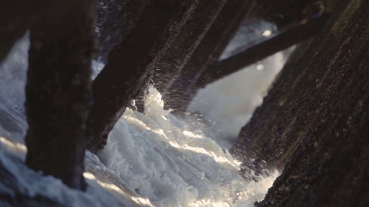 Waves crashing under a pier, old wooden pillings with sunbeams coming through