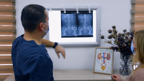 rear view of the doctor hanging x-ray scan on the light screen. medic points at organ image explaining the issue to a female patient.