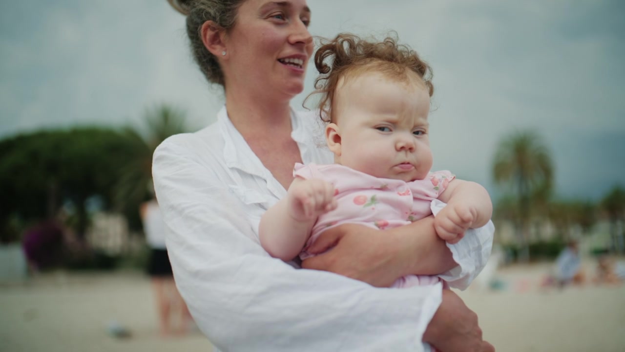 Neugieriges Baby lehnt sich vor, während es von einer Mutter nahe der Küstenlinie gehalten wird, und beobachtet sanfte Meereswellen während eines Familienstrandurlaubs.