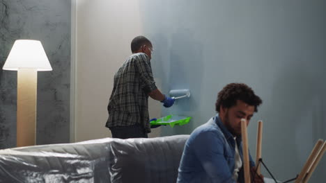 black man applies paint on wall friend fixes chair in room