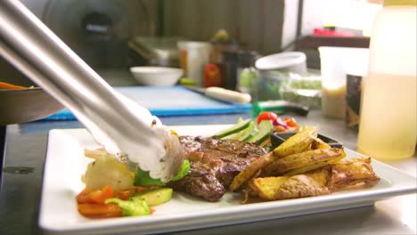 chef using metal tongs to place grilled vegetables on plate with steak and potatoes