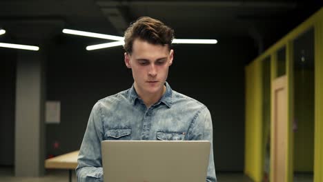 un joven atractivo con camisa de mezclilla con una laptop en las manos está caminando en algún lugar interior bien iluminado, totalmente concentrado