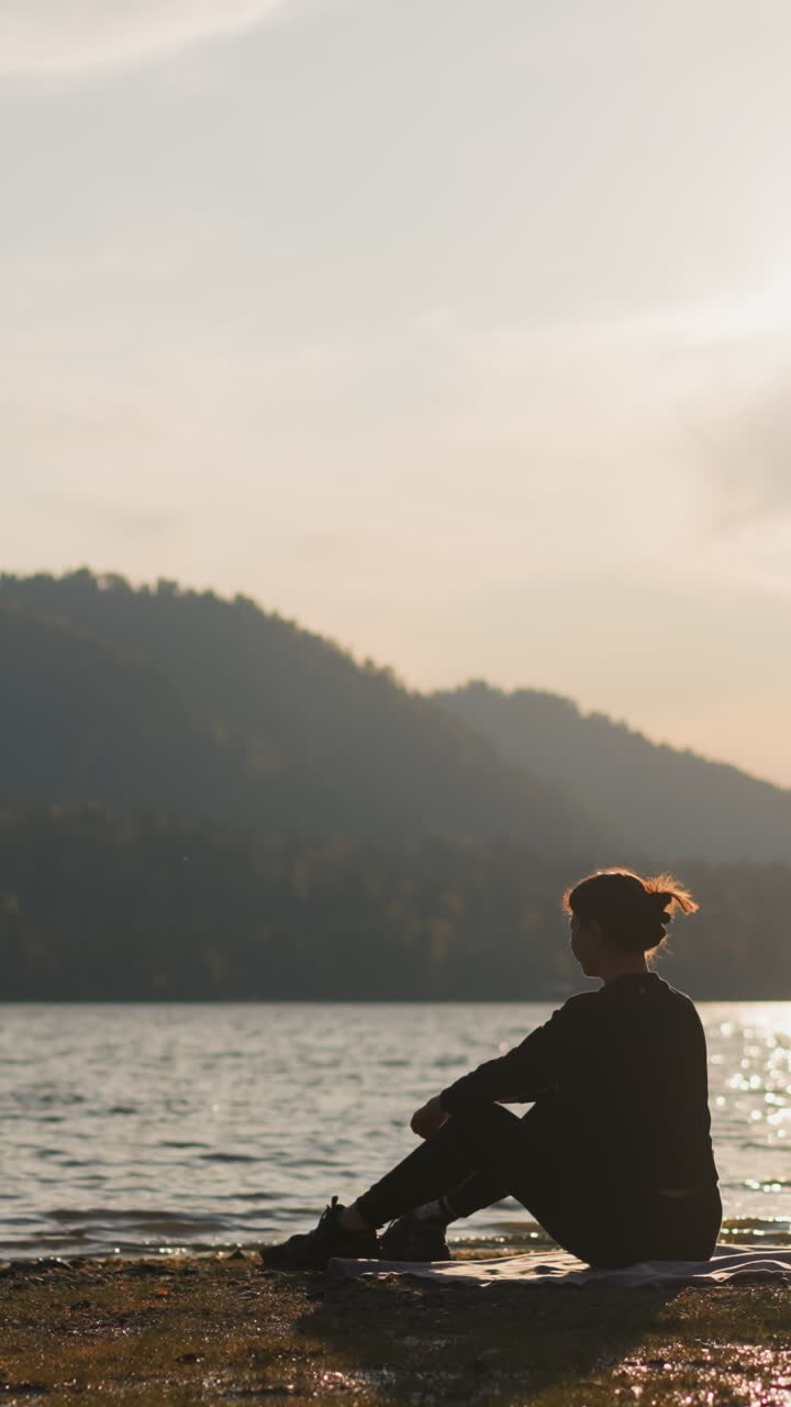 Woman throws rock into water of lake at sunset. Lady silhouette enjoys picnic on riverbank alone with nature surrounded by evening view of autumn mountains