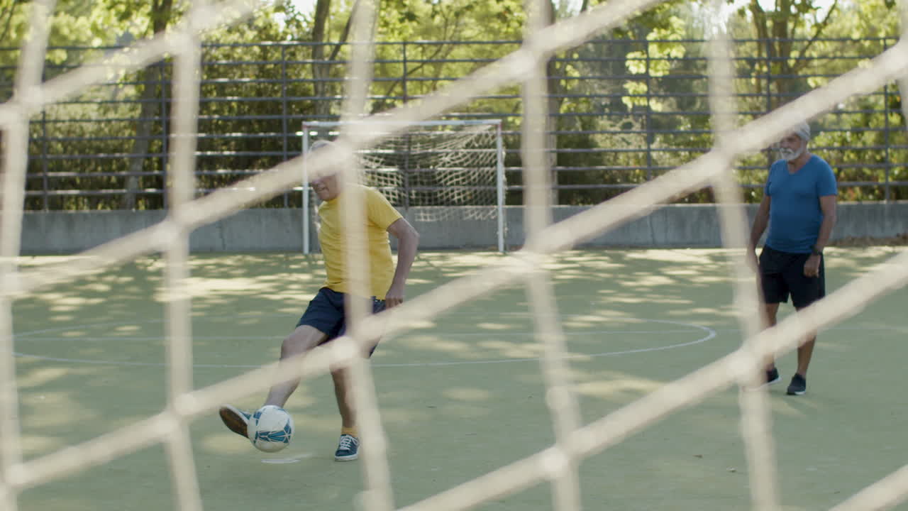 vista frontal de un anciano feliz pateando la pelota, anotando un gol y celebrando mientras juega fútbol con amigos en un estadio
