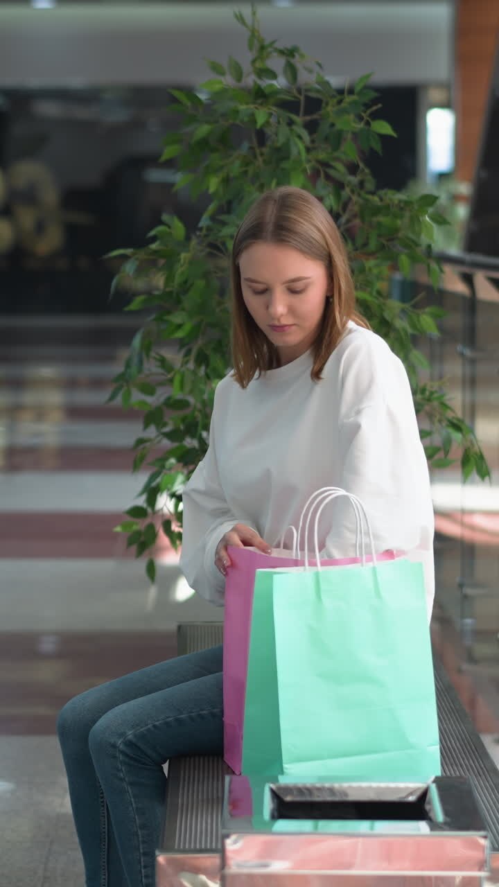 Female shopper seated in bright modern shopping mall retrieves yellow-covered book from pink bag, casually opening it while surrounded by green plants, glass railings, and spacious reflective floors