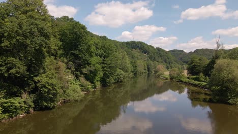 The-Sieg-flowing-calmly-in-between-lush,-leafy-riverbanks-underneath-a-blue-sky-with-cumulus-clouds