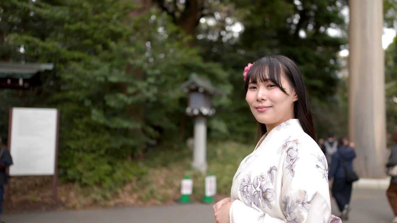 Una mujer japonesa sonriente con un kimono floral blanco de pie frente a una gran puerta torii