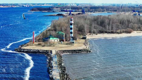 A tall black and white striped lighthouse rises on a sandy peninsula lined with trees and buildings, dividing calm blue waterways as waves gently curve around the shoreline under clear skies.